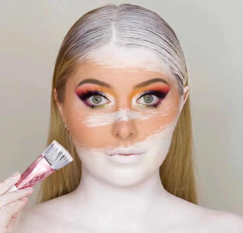 Paula Wolf mit buntem Augen-Make-up, halb weiß geschminktem Gesicht und Pinsel in der Hand